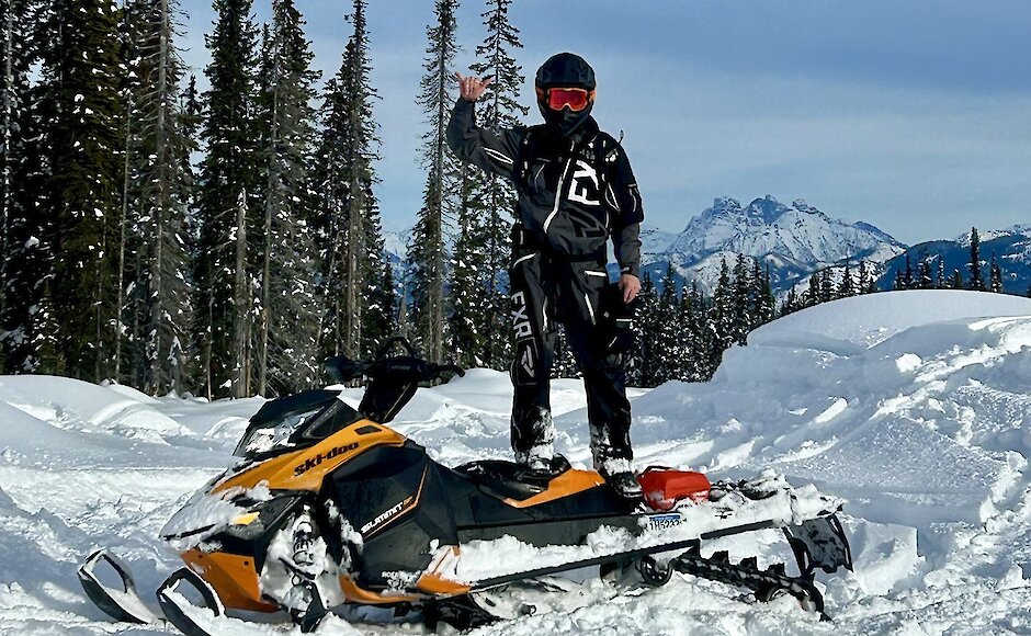 Snowmobile rider standing on their sled and giving the "hang loose" sign with snowy forest and BC mountains in the background.