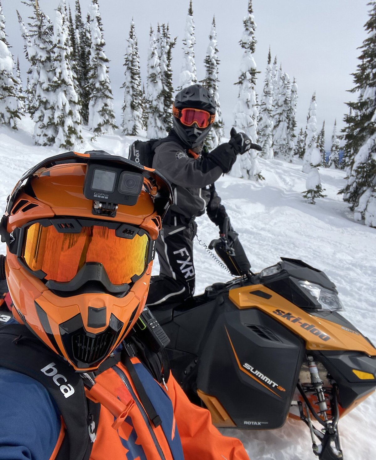 Snowmobile riders on their sleds with Kamloops Snowmobile Adventures and giving the "hang loose" sign with snowy forest in the background.