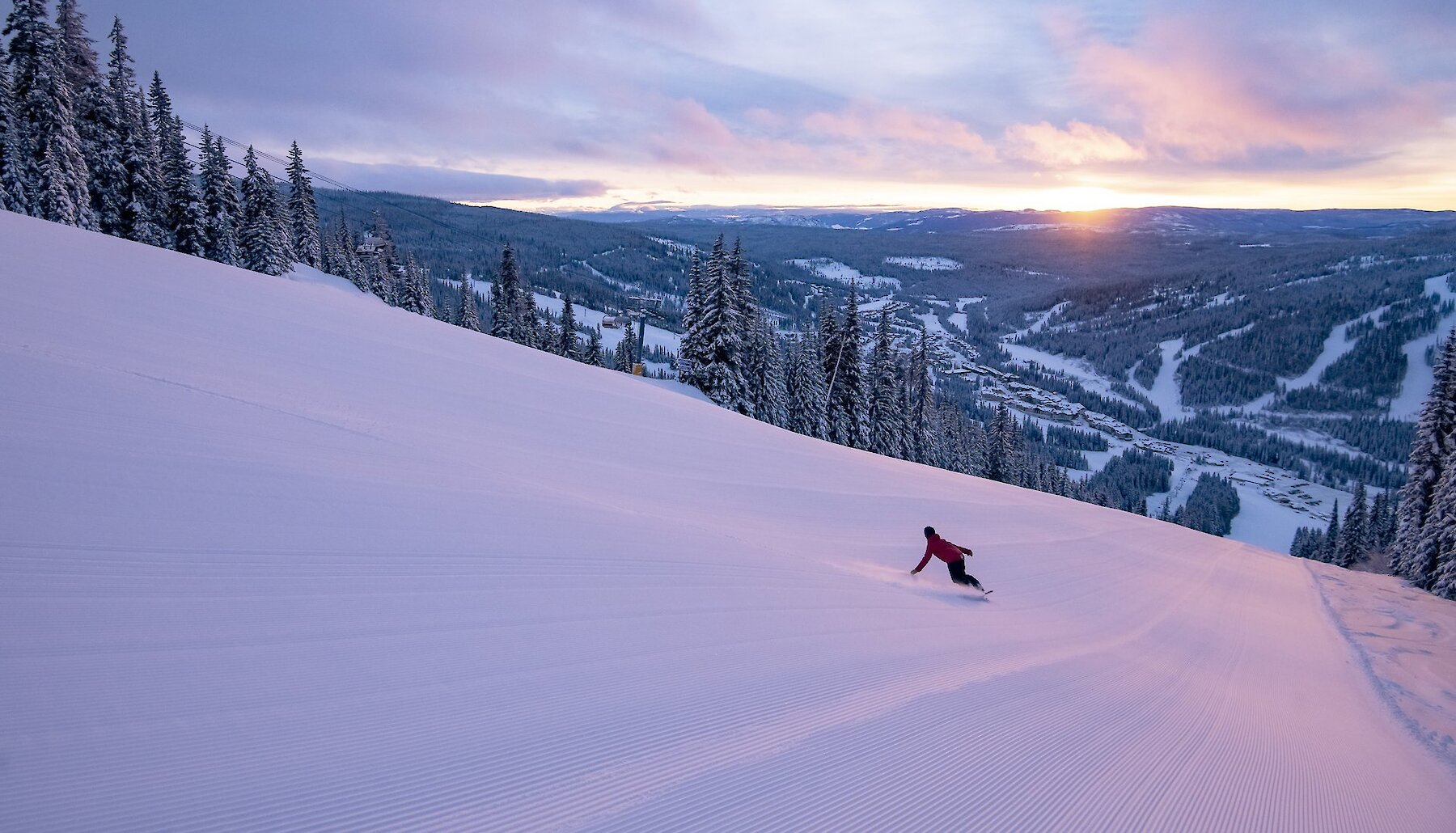 A snowboarder carves down an open, freshly groomed run at Sun Peak BC as the sun sets in the distance, casting gold and purple light over the scene.