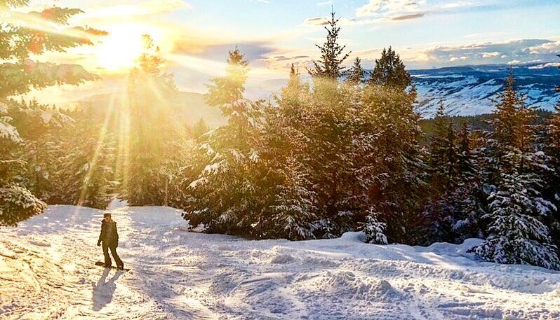 A snowboarder pauses on a snowy trail at Harper Mountain near Kamloops BC, under a blue sky and bright sunlight.