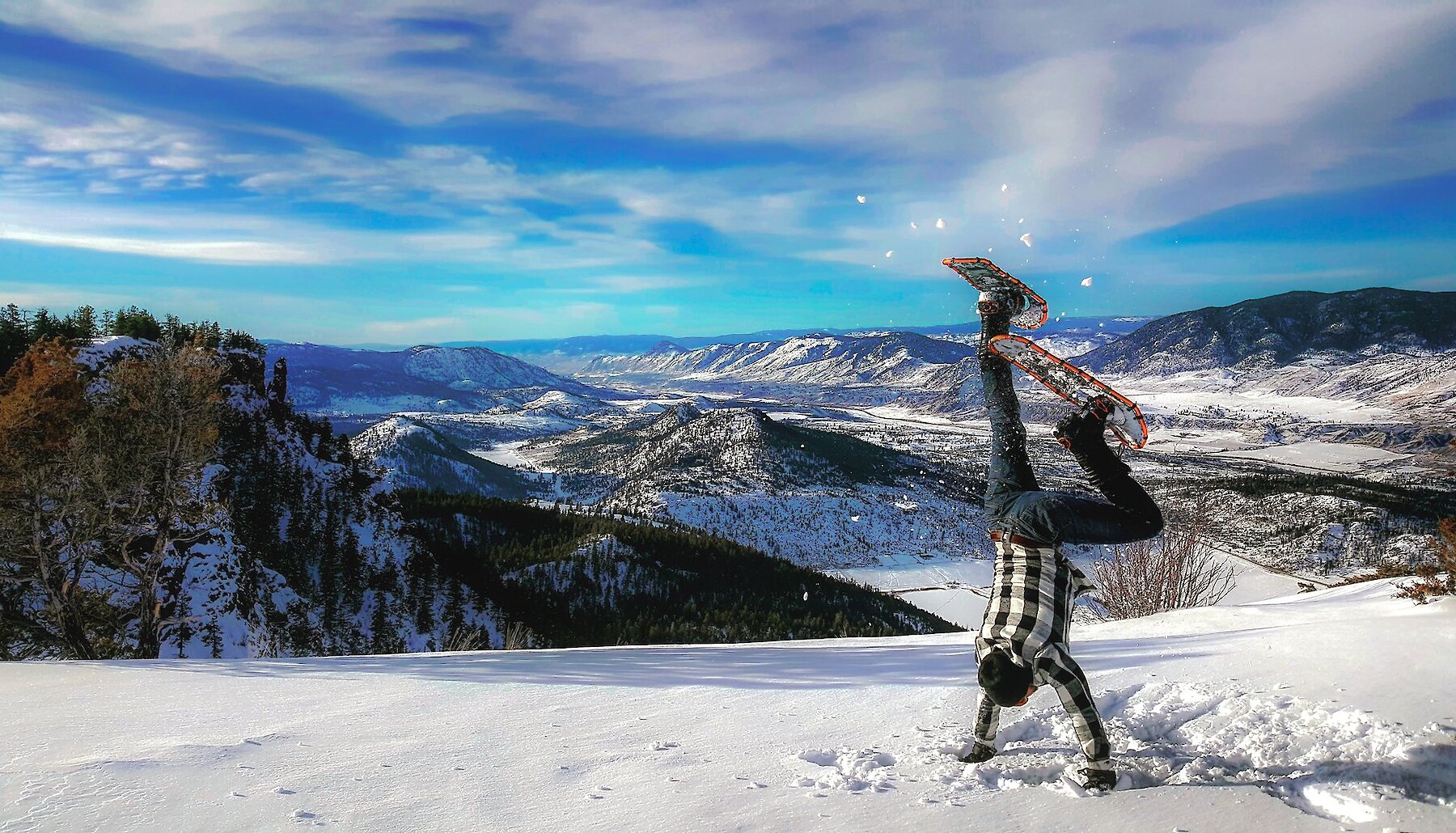 A person in snowshoes does a handstand in the snow on top of Buse Hill, overlooking the valley towards Kamloops BC.