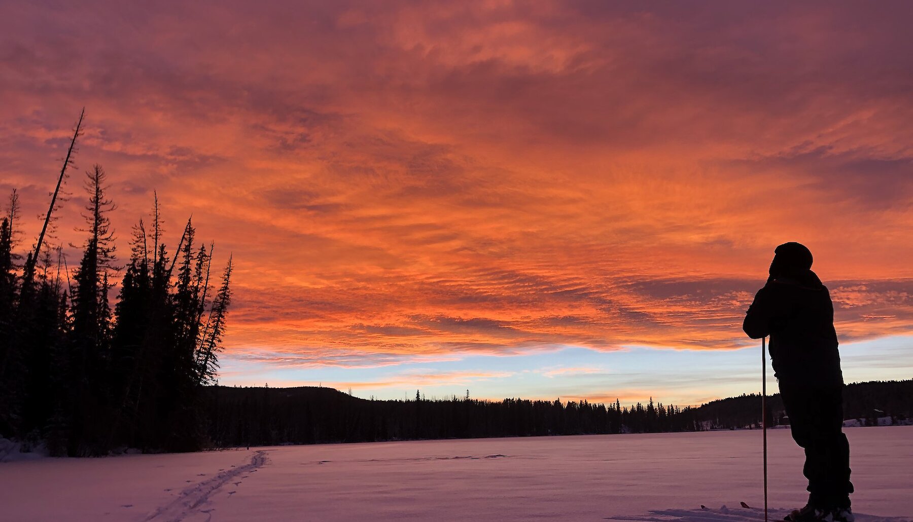 A person in cross-country skis stands on the ice at Lac le Jeune, near Kamloops BC, watching a dramatic gold and purple sunset.