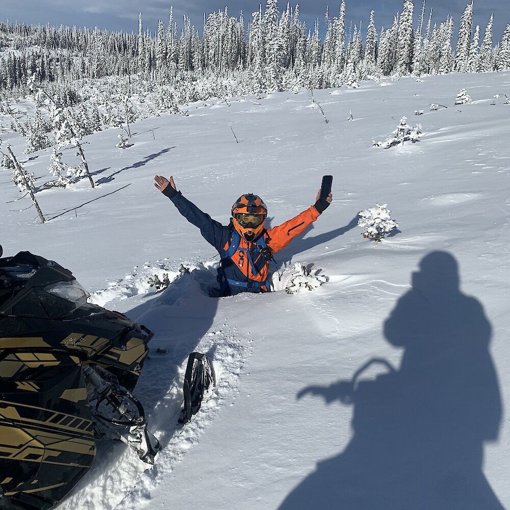 A person is seen waist deep in snow next to a snowmobile in an open area with trees in the background.