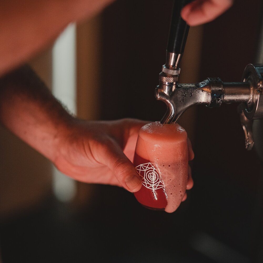 A person fills a beer glass with the Bright Eye Brewing Kamloops logo on it from a keg. The beer is red in colour, and foam overflows the edge.