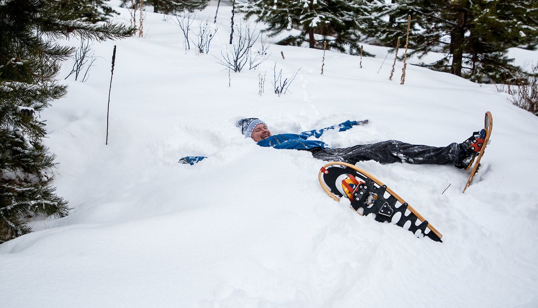 A person in snowshoes lies back in deep snow, smiling at the camera.