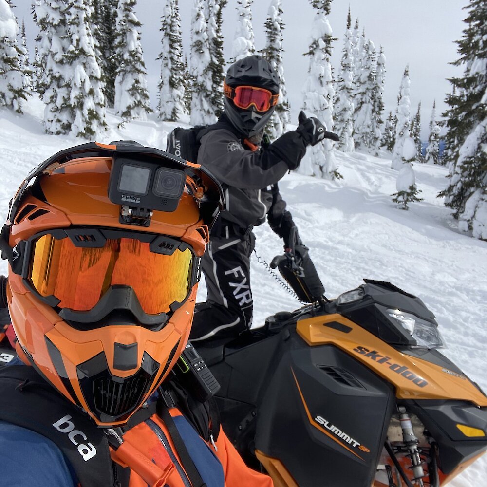 A couple snowmobilers geared up on their sleds giving the shaka sign as they ride a trail near Kamloops, BC.