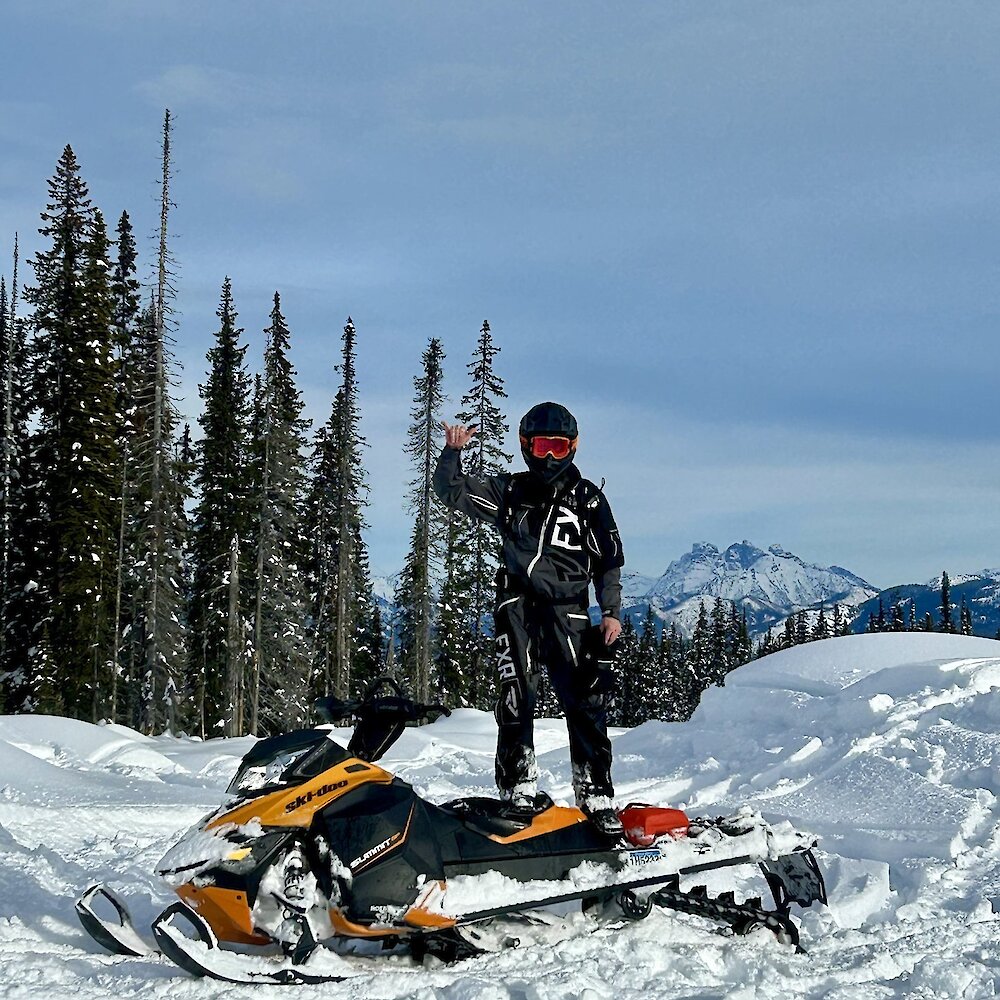 Snowmobiler standing on their sled giving the shaka sign with the snowy BC forest and mountains in the background.