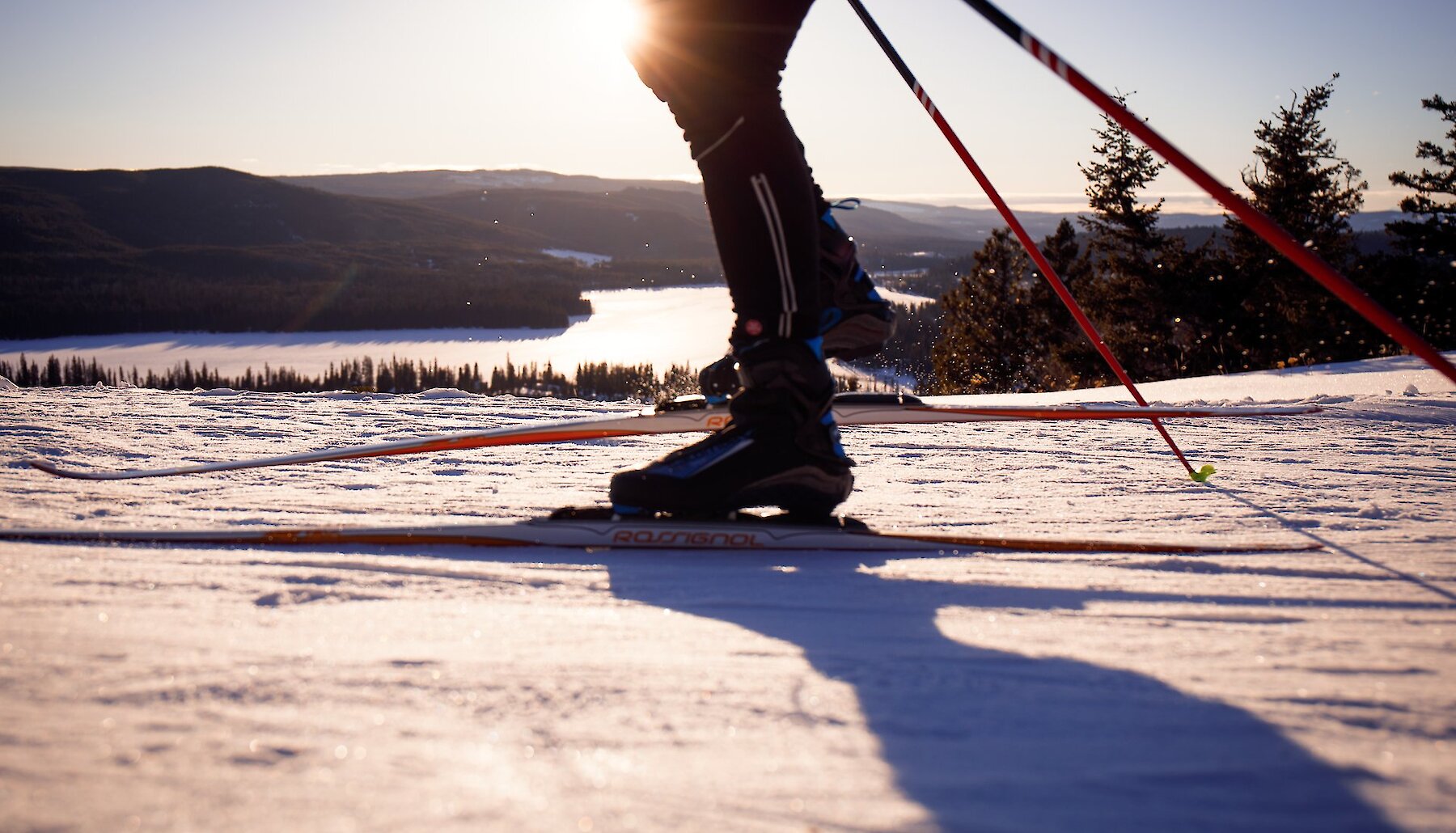 Cross country skier at Stake Lake Ski trail with the frozen lake, mountains, and sun in the background.