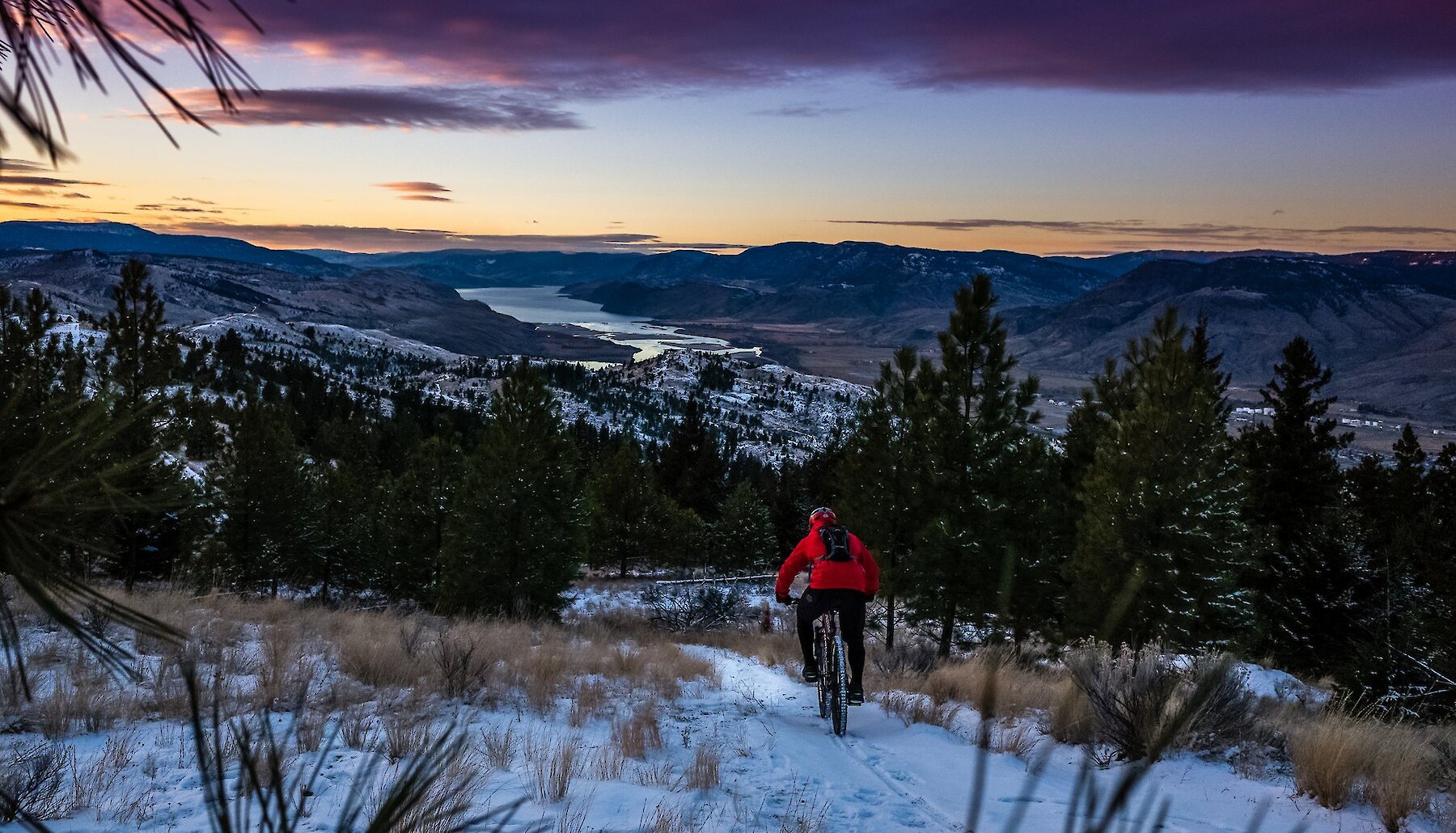 Fat biker rides down a snowy trail with the city of Kamloops in the background.