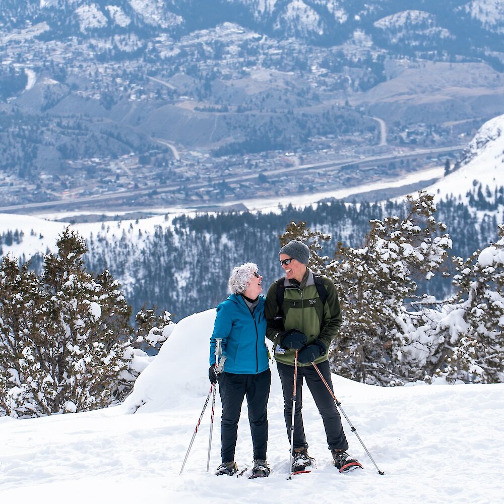 A couple smiling at each other wearing their snowshoes with the view from Harper Mountain lookout in the background.