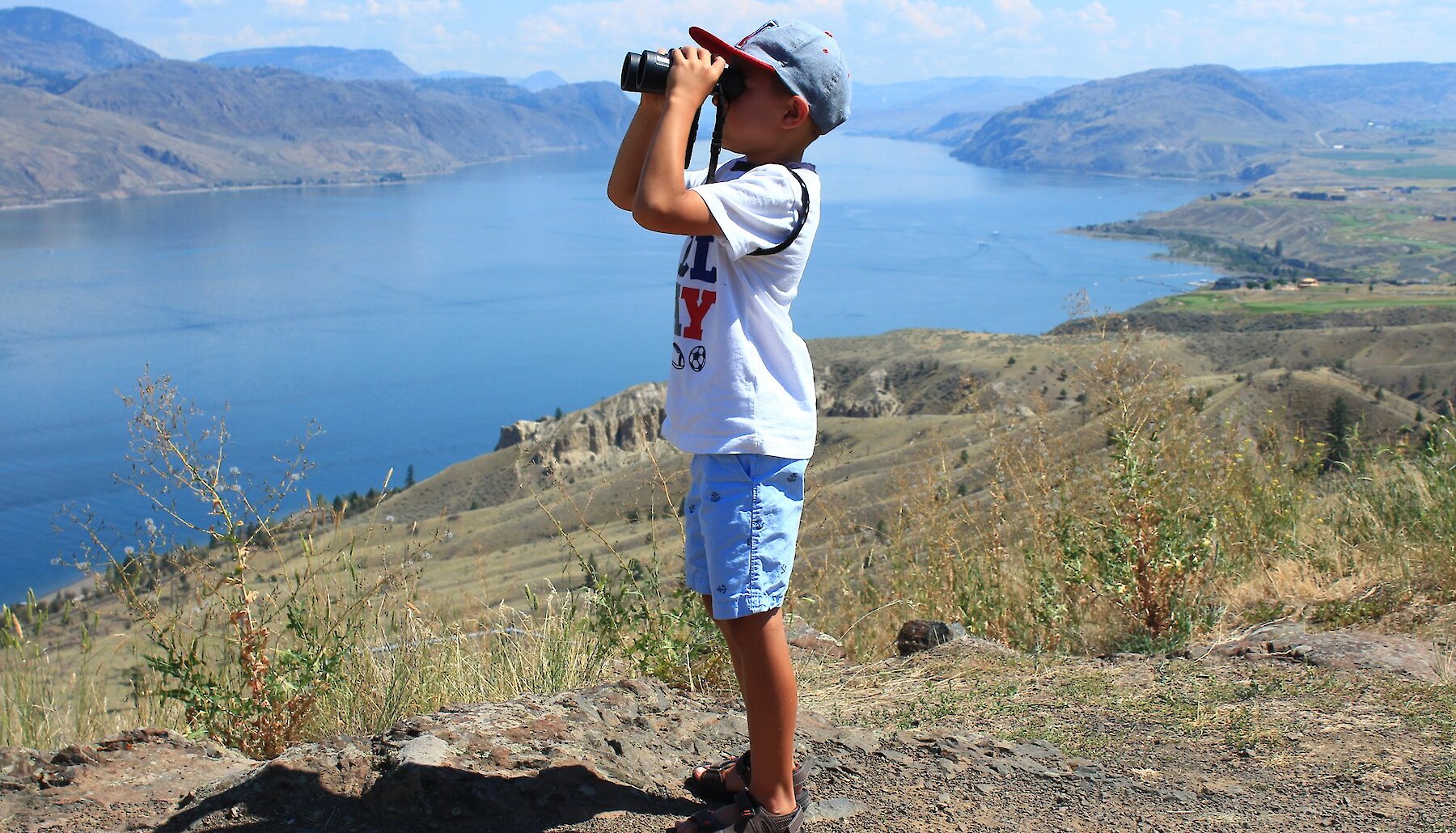 Boy looking through binoculars at the beautiful British Columbia scenery at a hiking viewpoint in Kamloops.