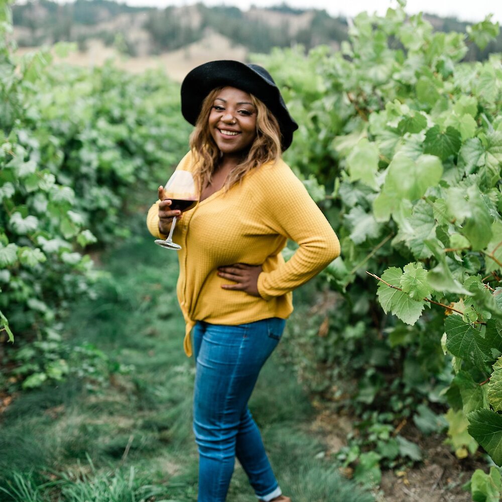 A woman stands in a vineyard holding a glass of wine while she smiles at the camera.