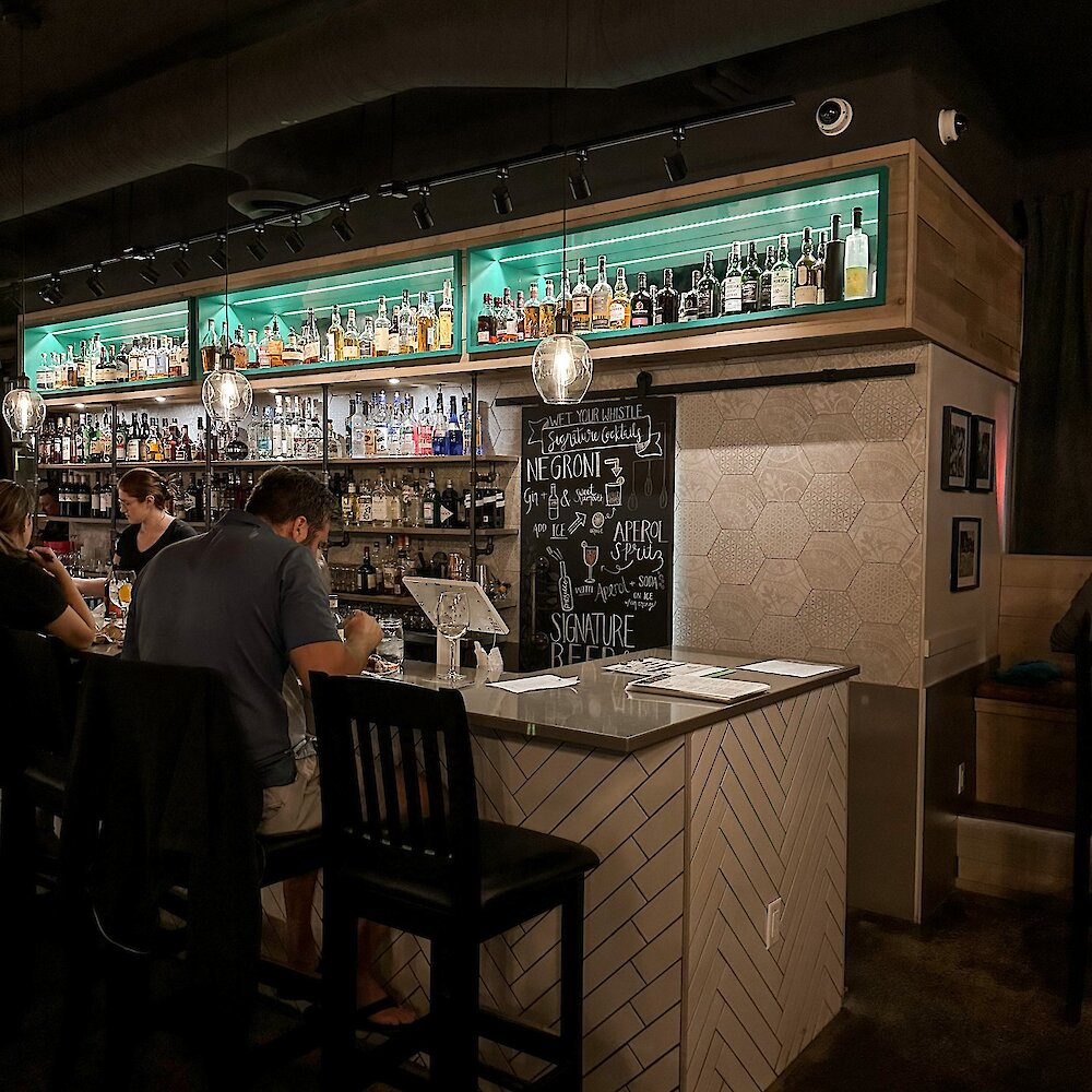 People sit on stools at a bar overlooking a brightly lit wall of spirits.