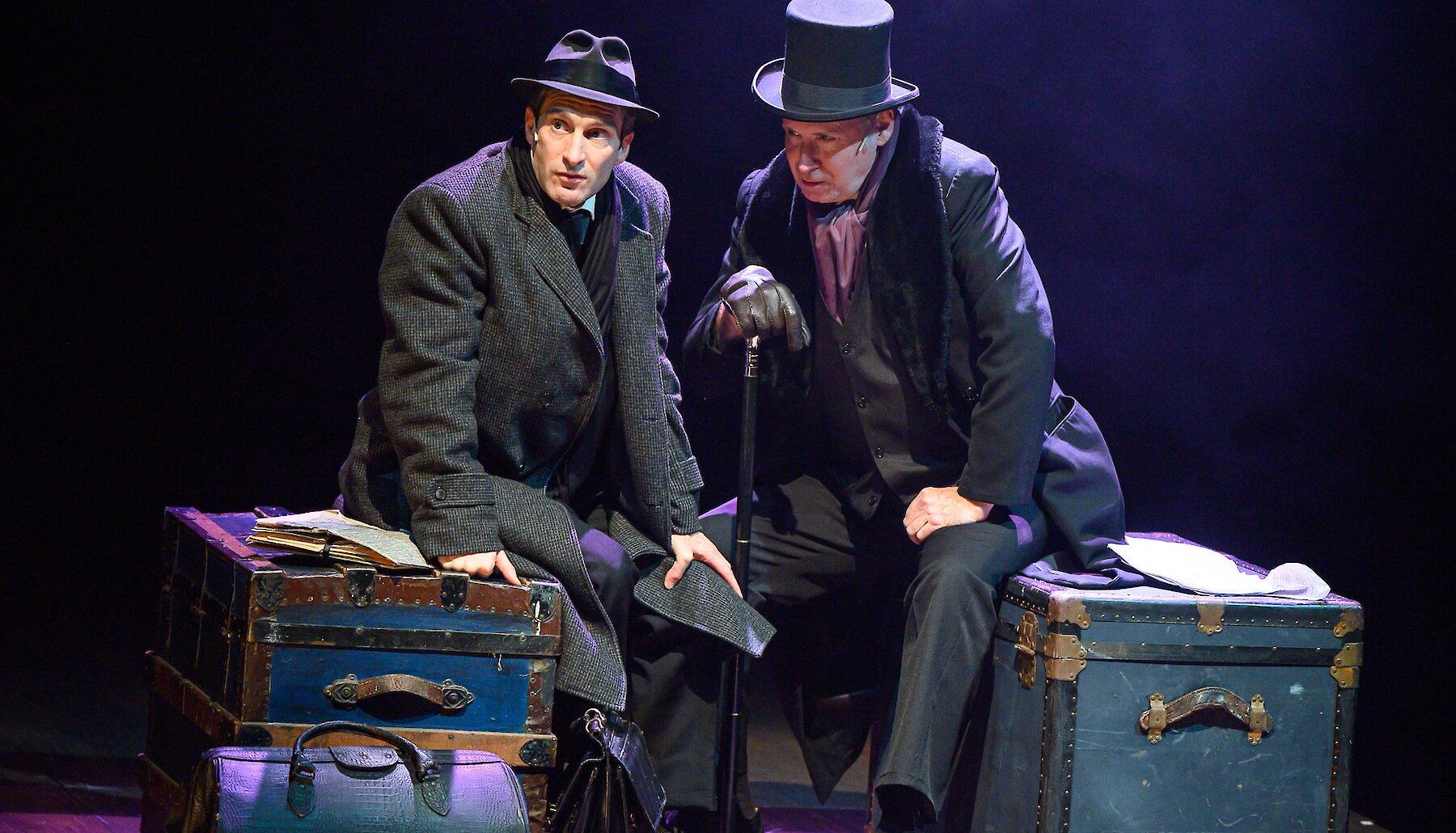 Two actors dressed in suits and trench coats sit onstage atop a pile of luggage.