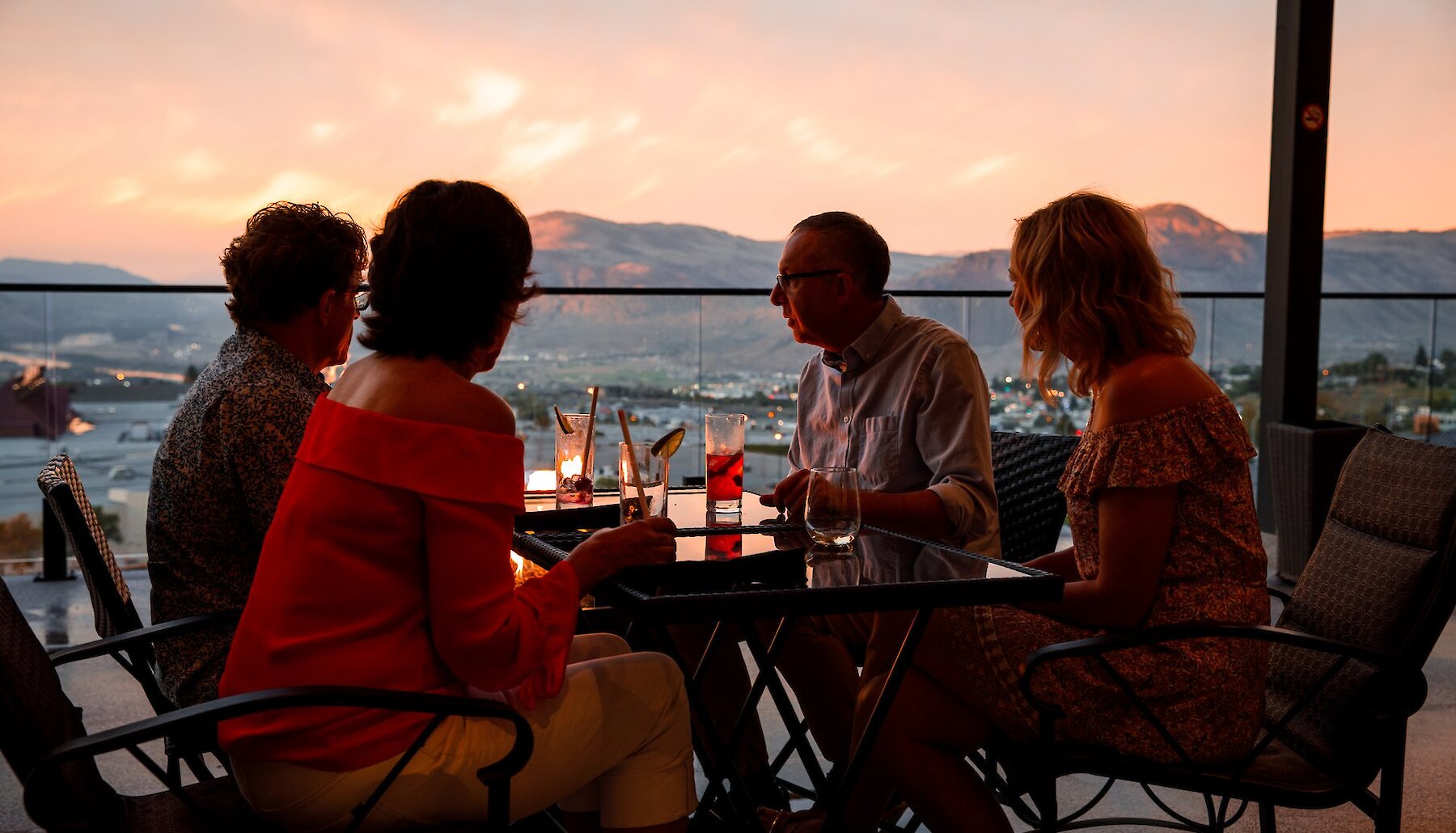 A group of four people dine outside on a patio overlooking the city at sunset.