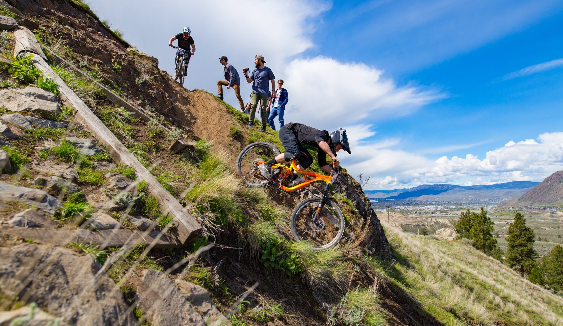 Two mountain bikers race down a steep hill as three friends cheer them on.