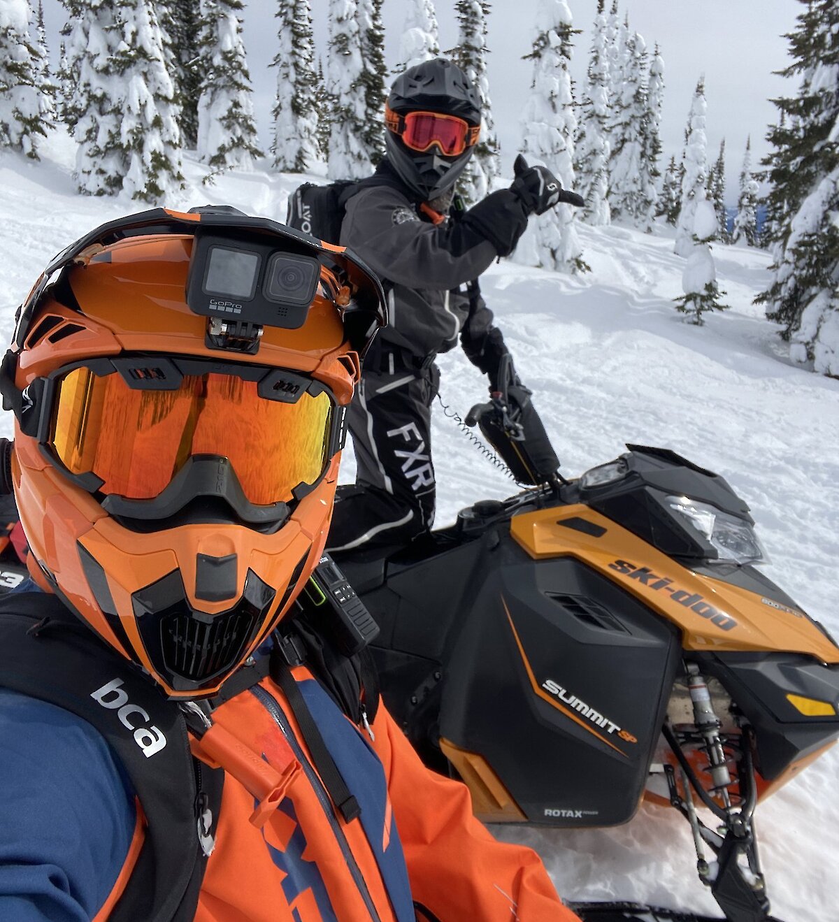 Snowmobile riders on their sleds with Kamloops Snowmobile Adventures and giving the "hang loose" sign with snowy forest in the background.